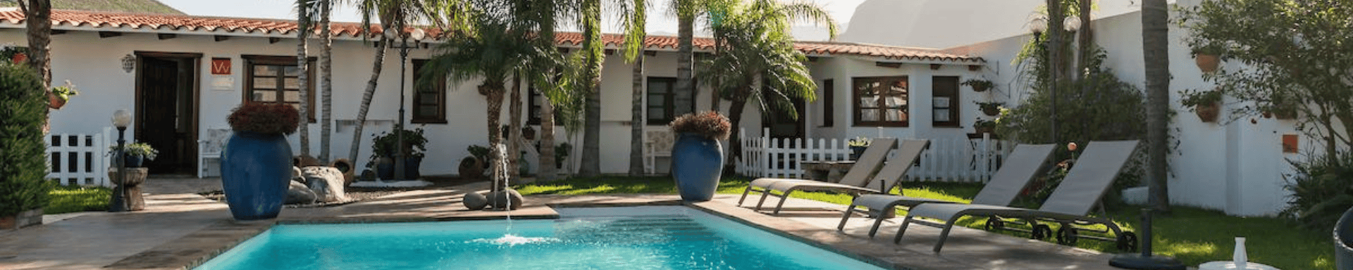 Vakantieverblijf met witgekalkte muren, een zwembad, ligstoelen en tropische planten.