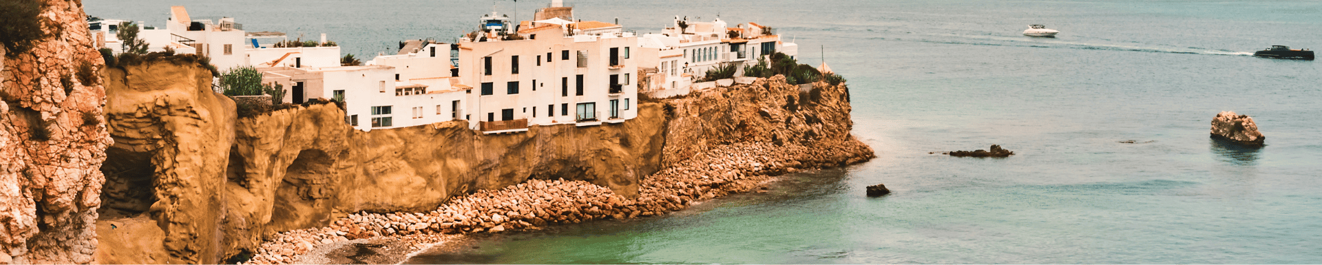 Witgekalkte gebouwen op een klif aan de kust van Spanje, met helderblauw water en boten op de achtergrond.